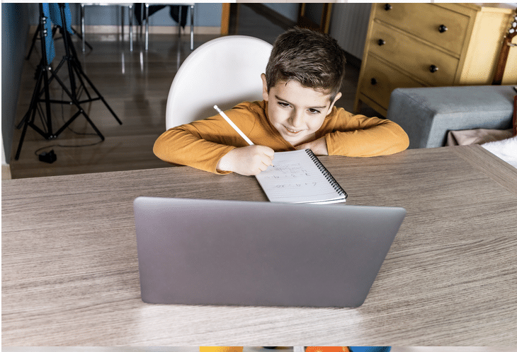 Child leaning forward on to desk, writing on paper and looking at a computer screen